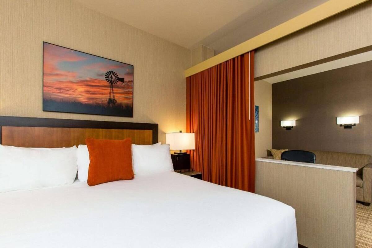 A hotel room with a bed, a desk, a chair, a nightstand, a lamp, a painting of a windmill, and a view of the city.