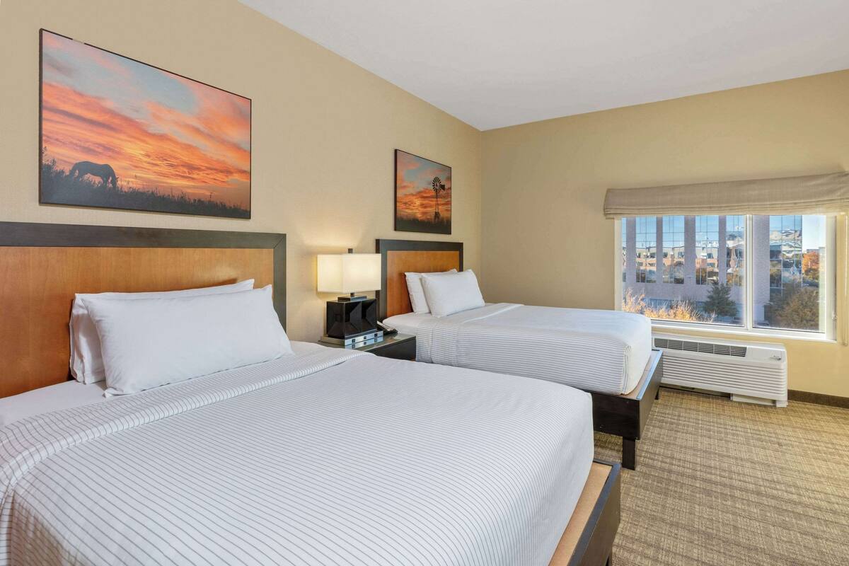 A hotel room with two beds, a nightstand with a lamp, a window with a view of buildings, and two paintings on the wall.
