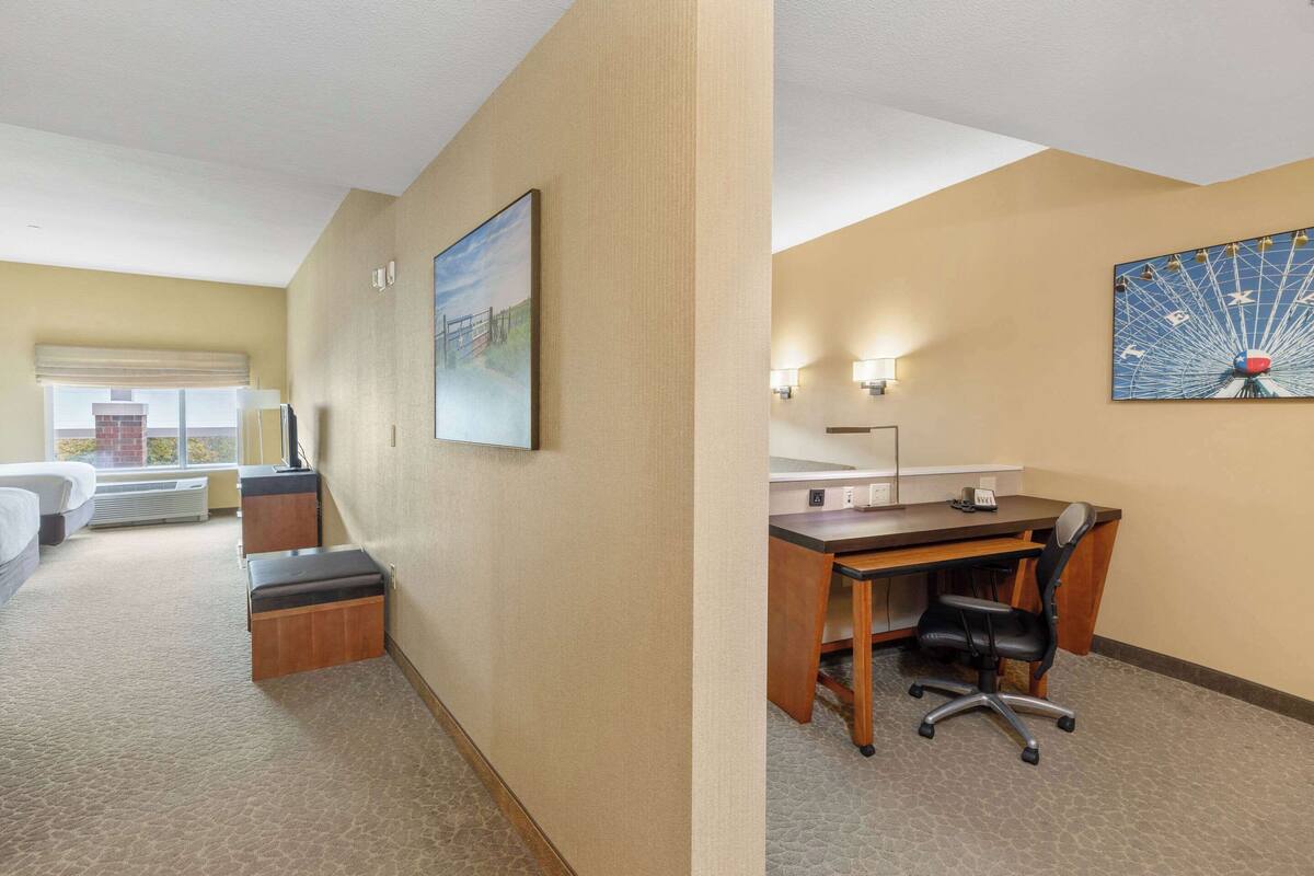 A hotel room with a bed, a desk, a chair, a painting, and a wall-mounted lamp.