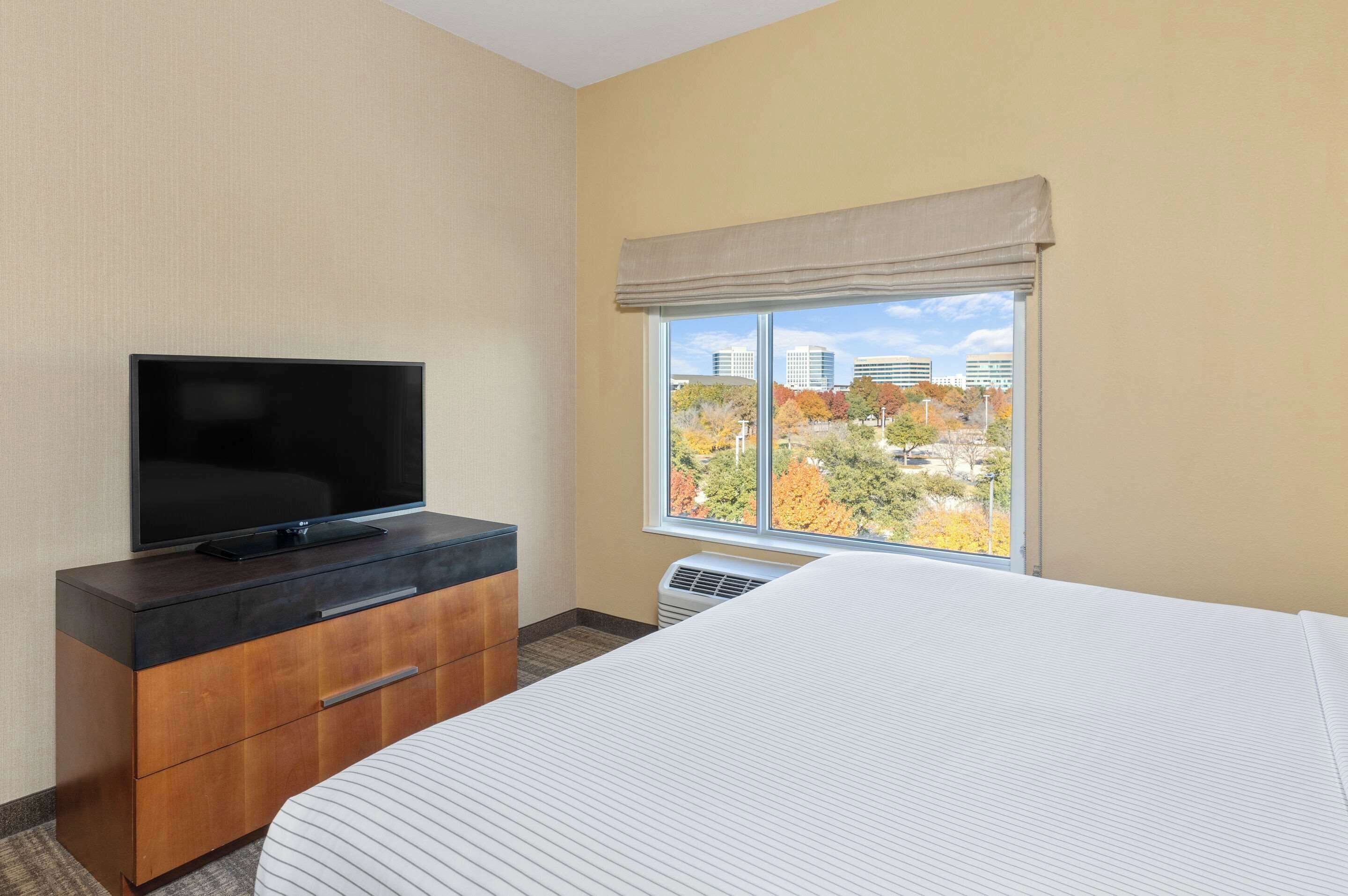 A hotel room with a bed, a TV on a wooden dresser, and a window with a view of autumn trees and buildings.