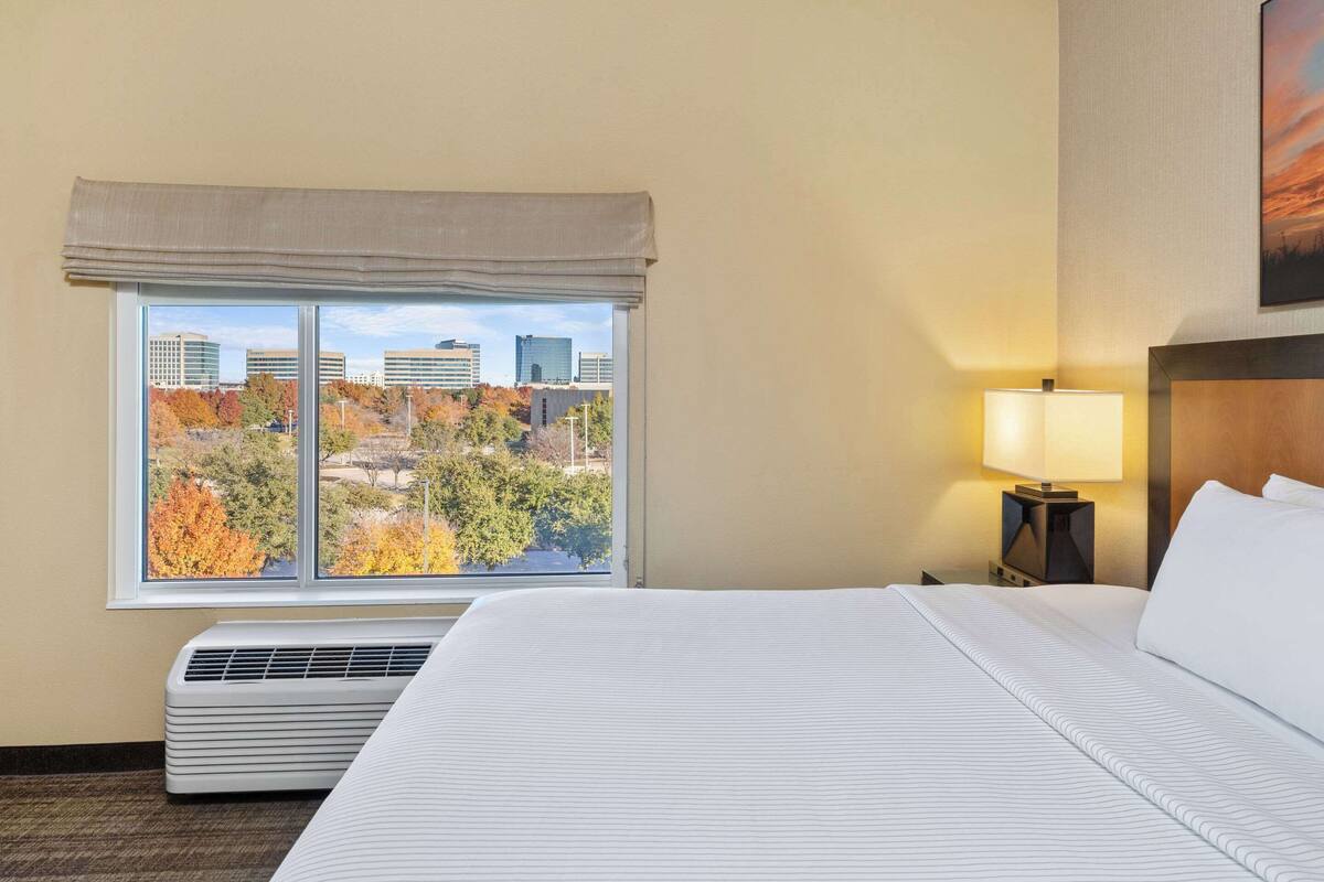 A hotel room with a large bed, a window with a view of buildings and autumn trees, a bedside lamp, and an air conditioning unit.