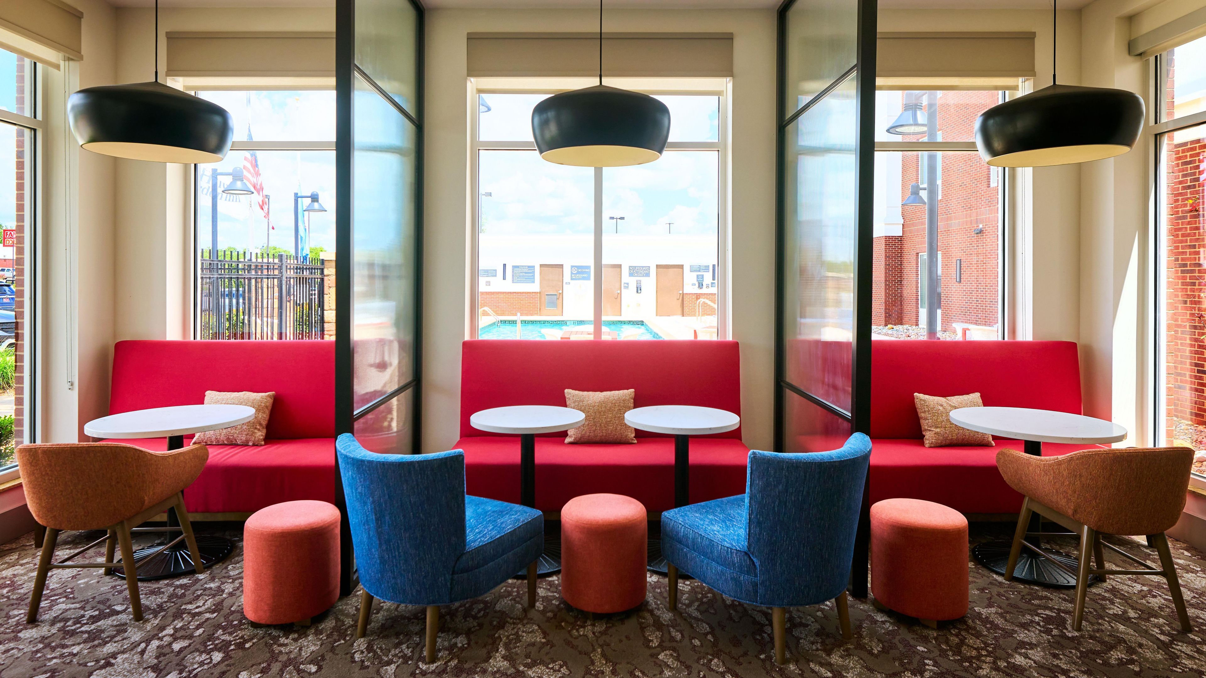 A seating area with red couches, blue chairs, and round tables, featuring large windows and hanging pendant lights.