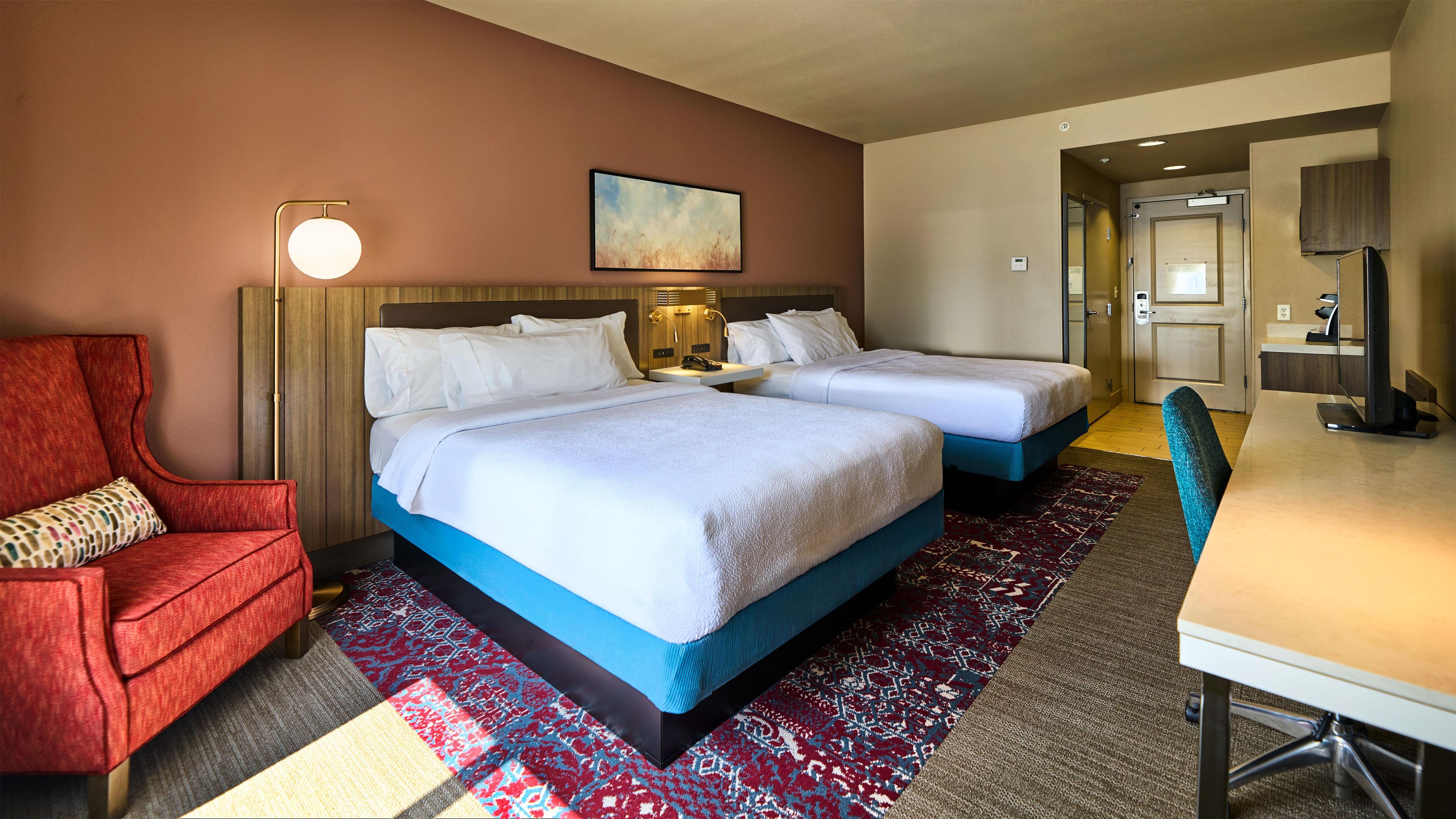 A hotel room with two beds, a desk, a chair, a lamp, and a painting on the wall.