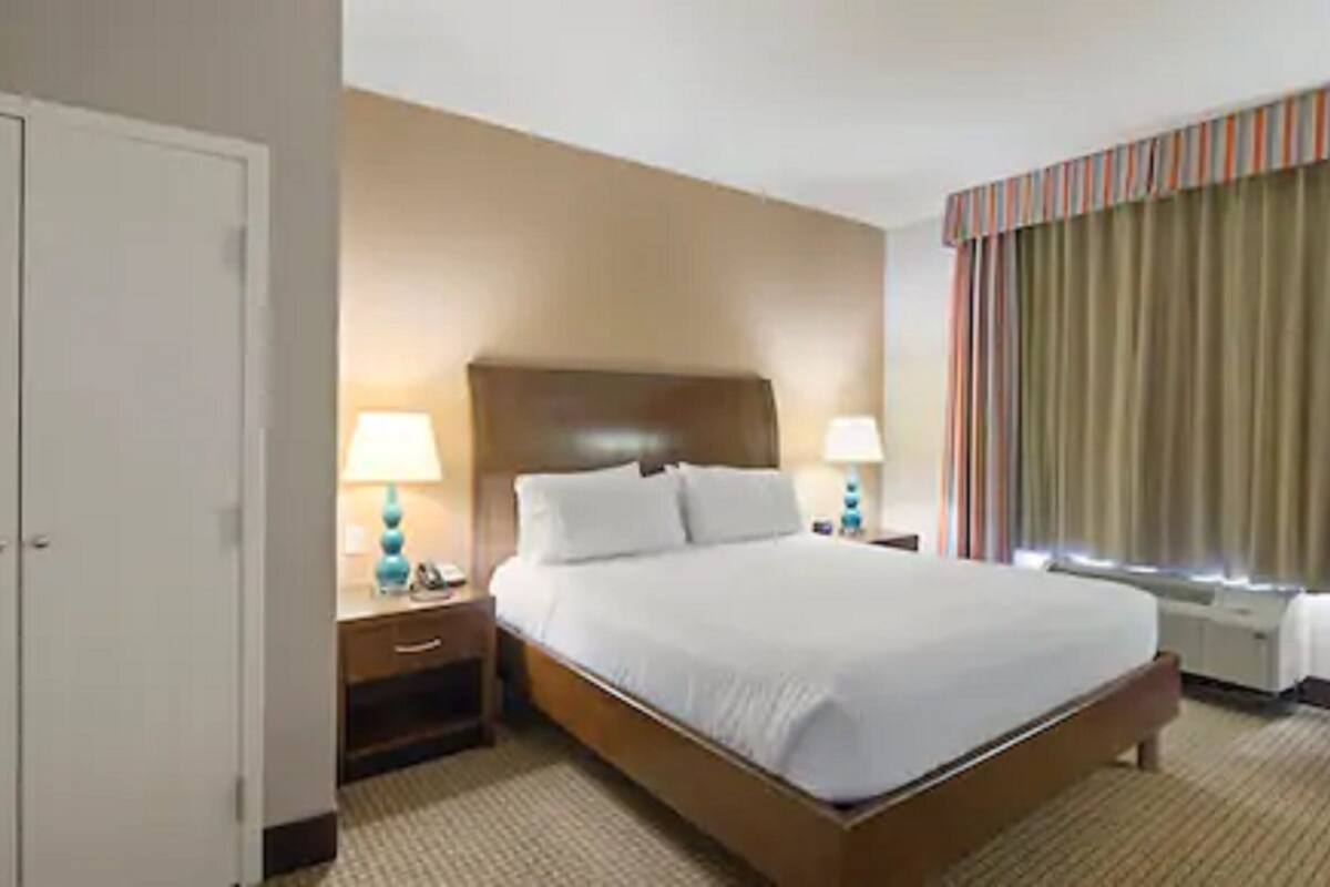 Habitación de hotel con una cama grande, dos mesitas de noche con lámparas, un armario y una ventana con cortinas.