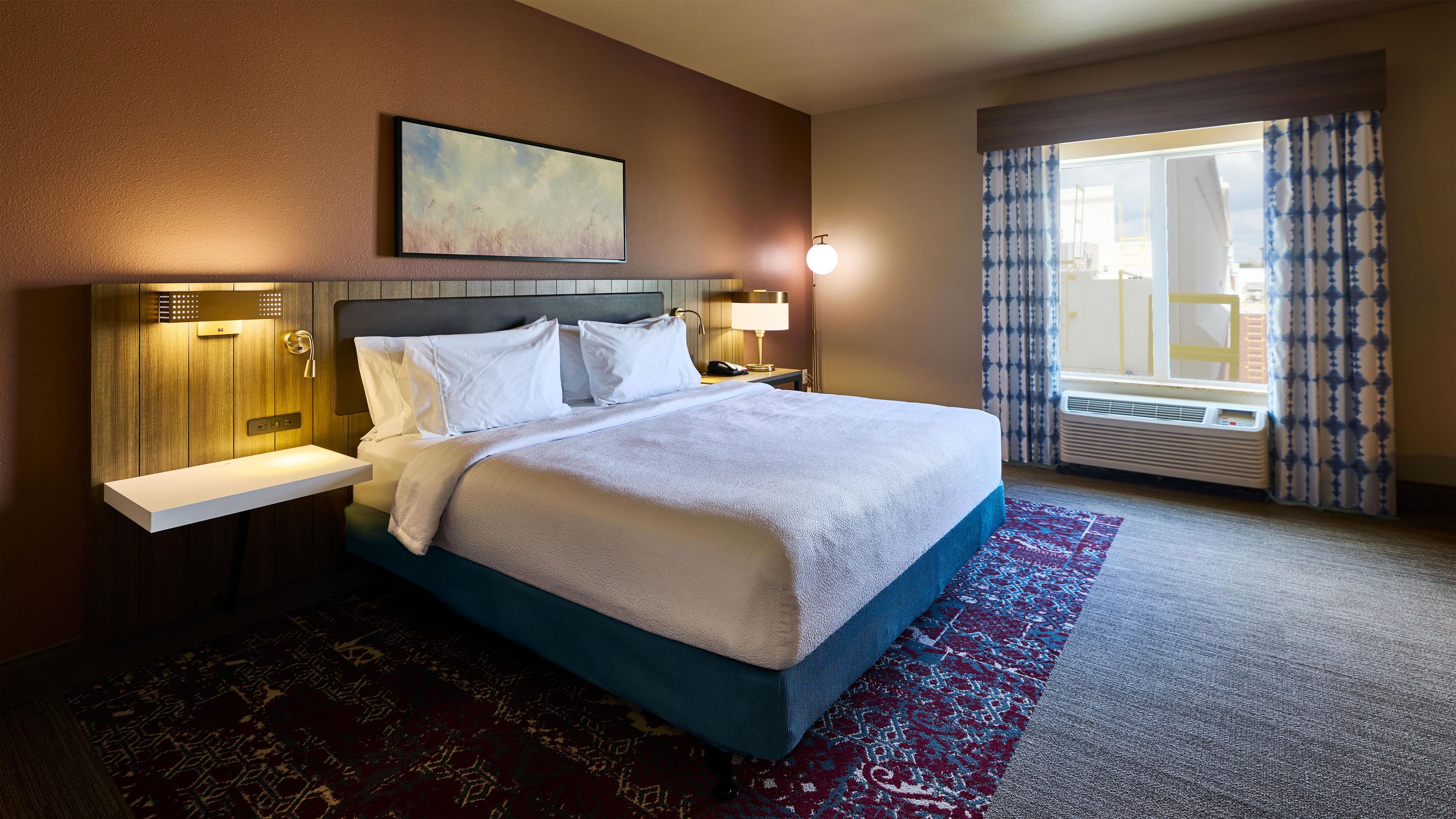 A hotel room with a large bed, bedside tables, a wall-mounted lamp, and a window with curtains.