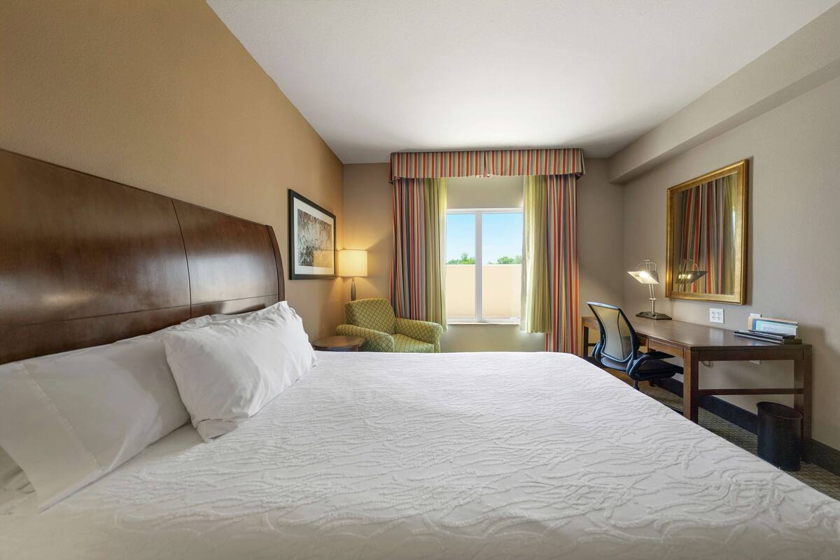 Una habitación de hotel con una cama grande, un escritorio con una silla, una lámpara y un ventanal con cortinas.