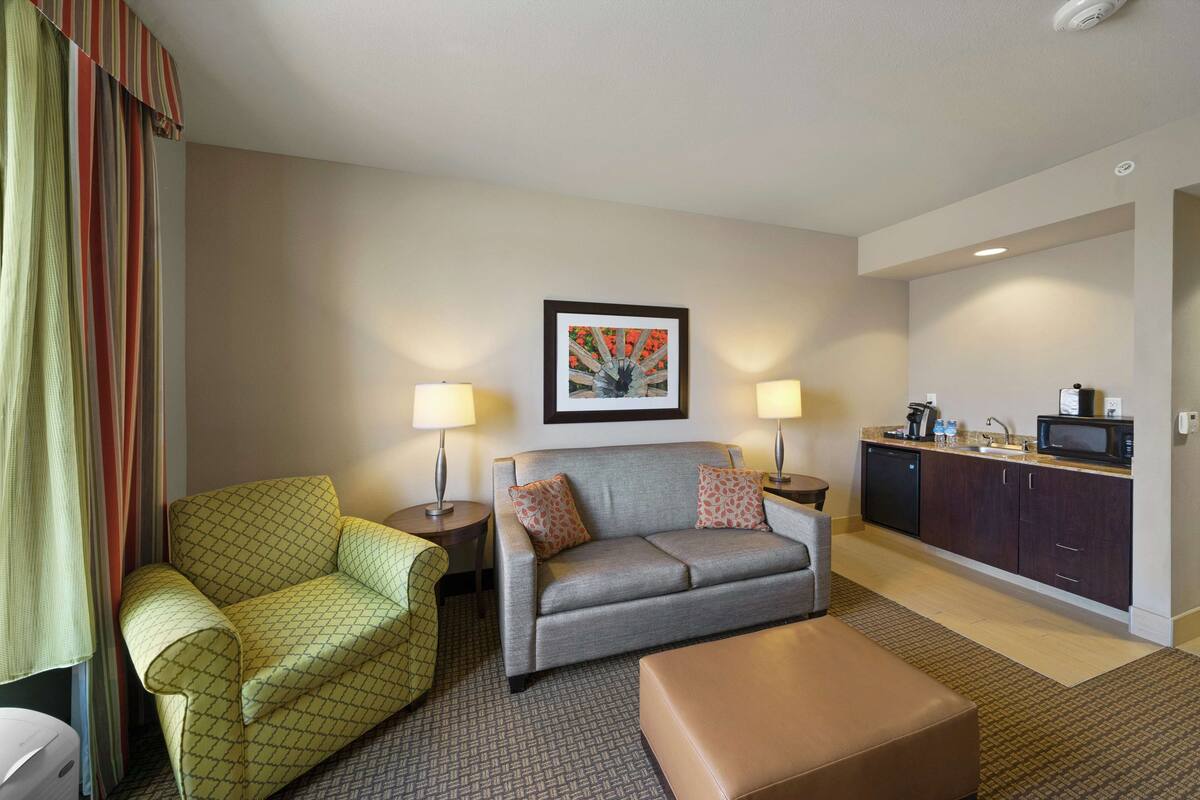 Una habitación de hotel con sofá, sillón y reposapiés, una kitchenette y un cuadro en la pared.