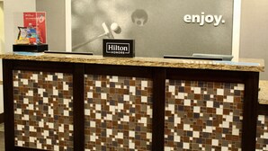 A reception desk with a mosaic tile design, a framed 'enjoy.' sign, and a 'Hilton Honors' sign. - Hampton Inn Indianapolis NW/Zionsville (Whitestown)