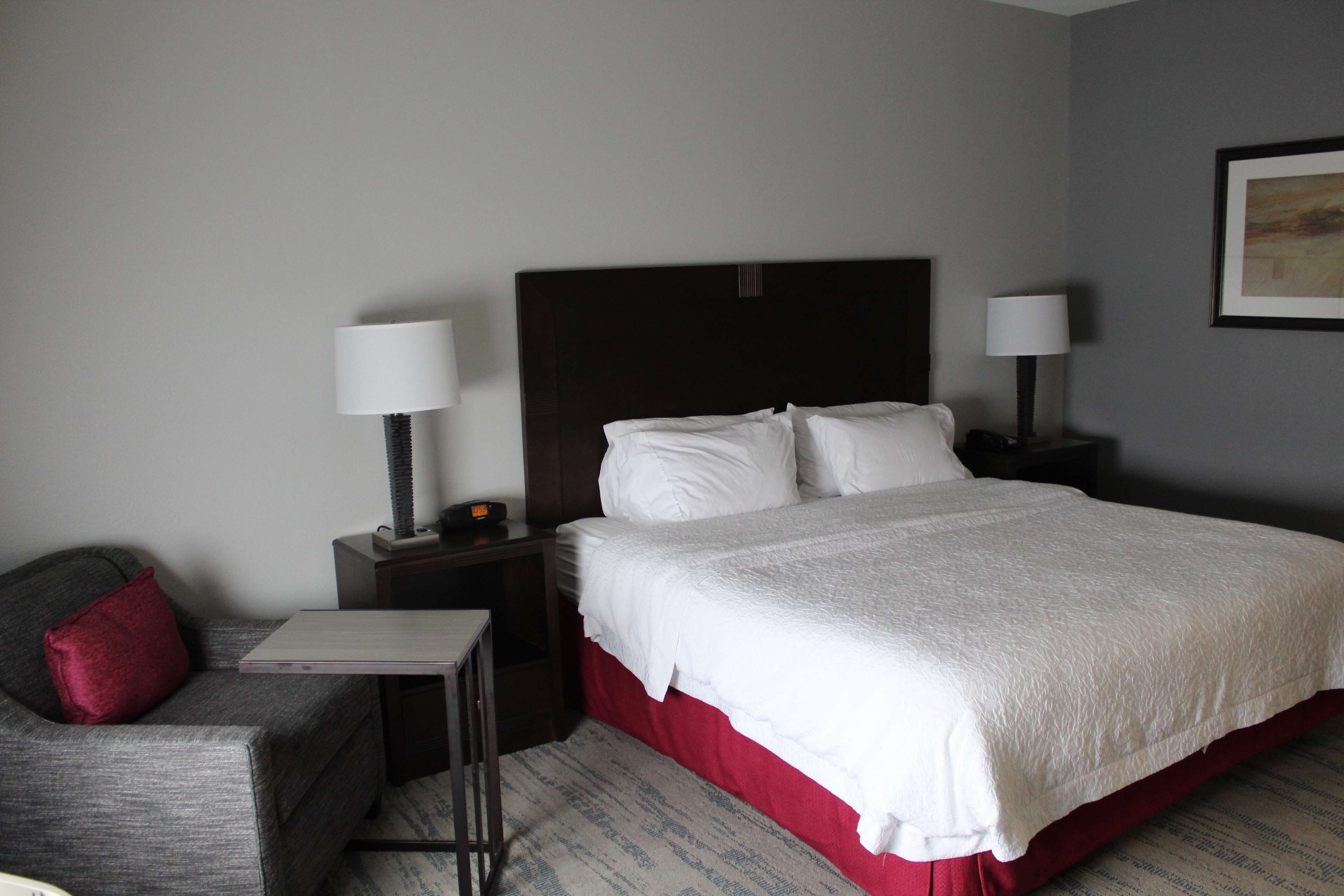 A hotel room with a bed, a nightstand, a lamp, a chair, and a small table.