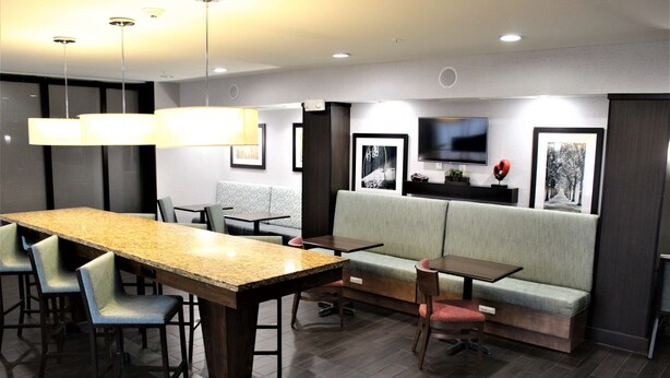 A modern hotel lobby with a long dining table, bar stools, a sofa, and a television mounted on the wall. - Hampton Inn Indianapolis NW/Zionsville (Whitestown)