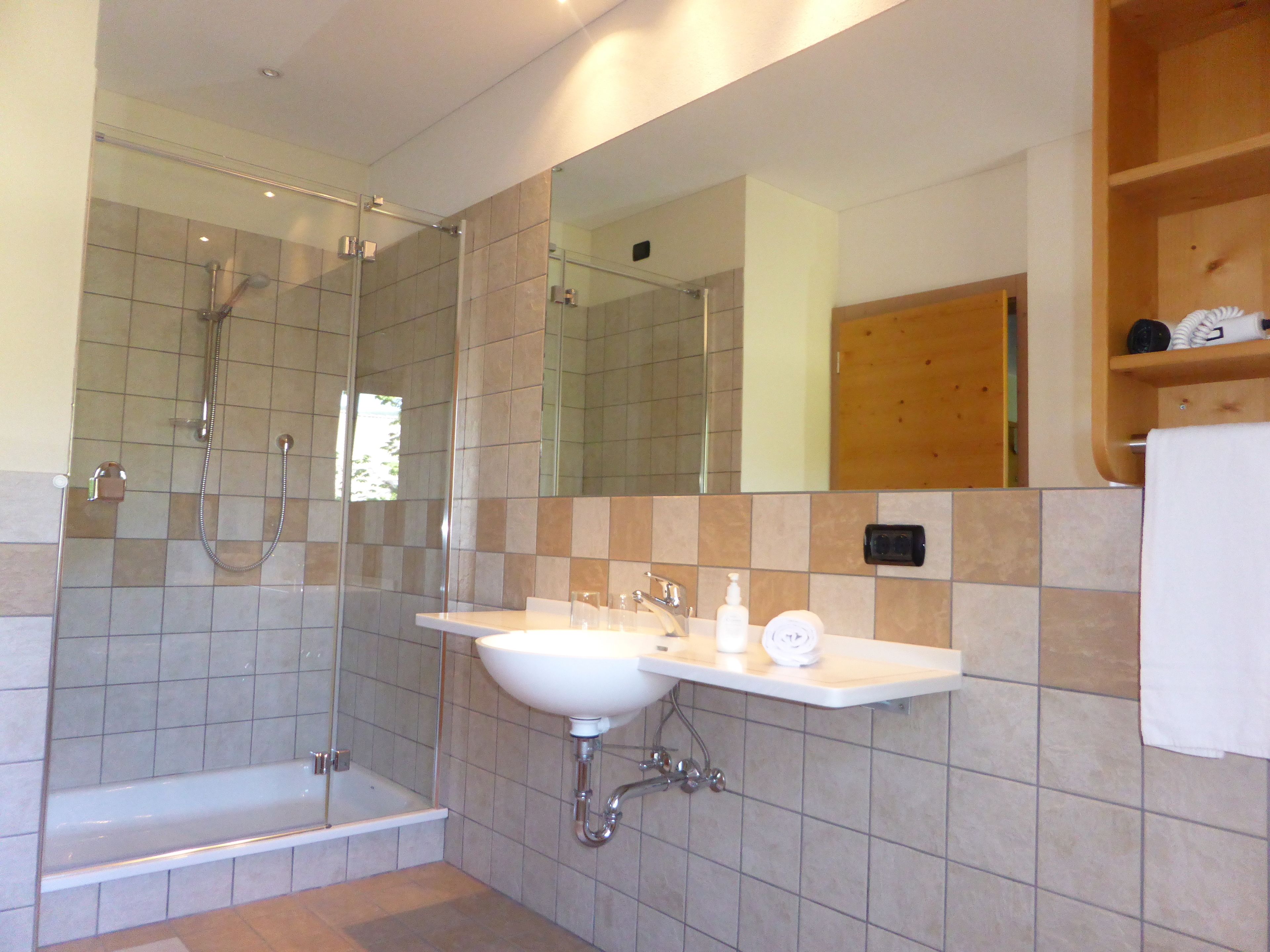 A modern bathroom with a glass-enclosed shower, a white sink, and a wooden shelf with toiletries.