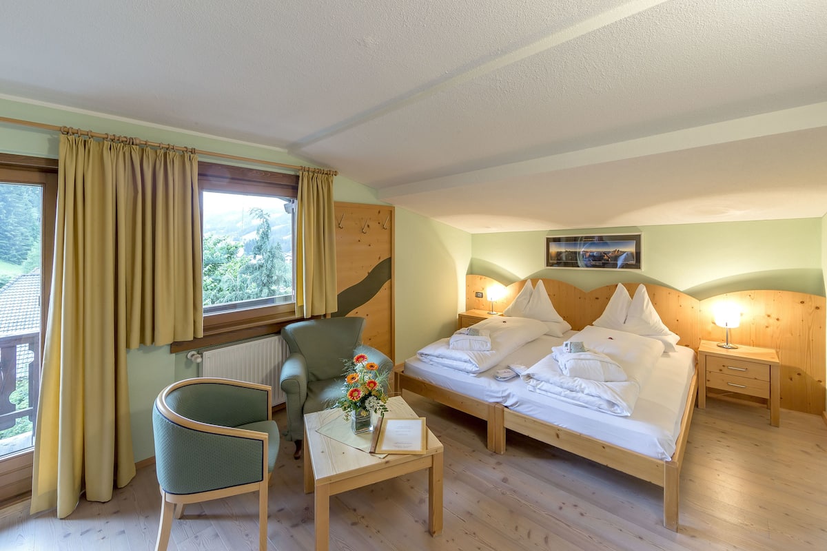 A room with a bed, a chair, a small table with a vase of flowers, and a window with curtains.