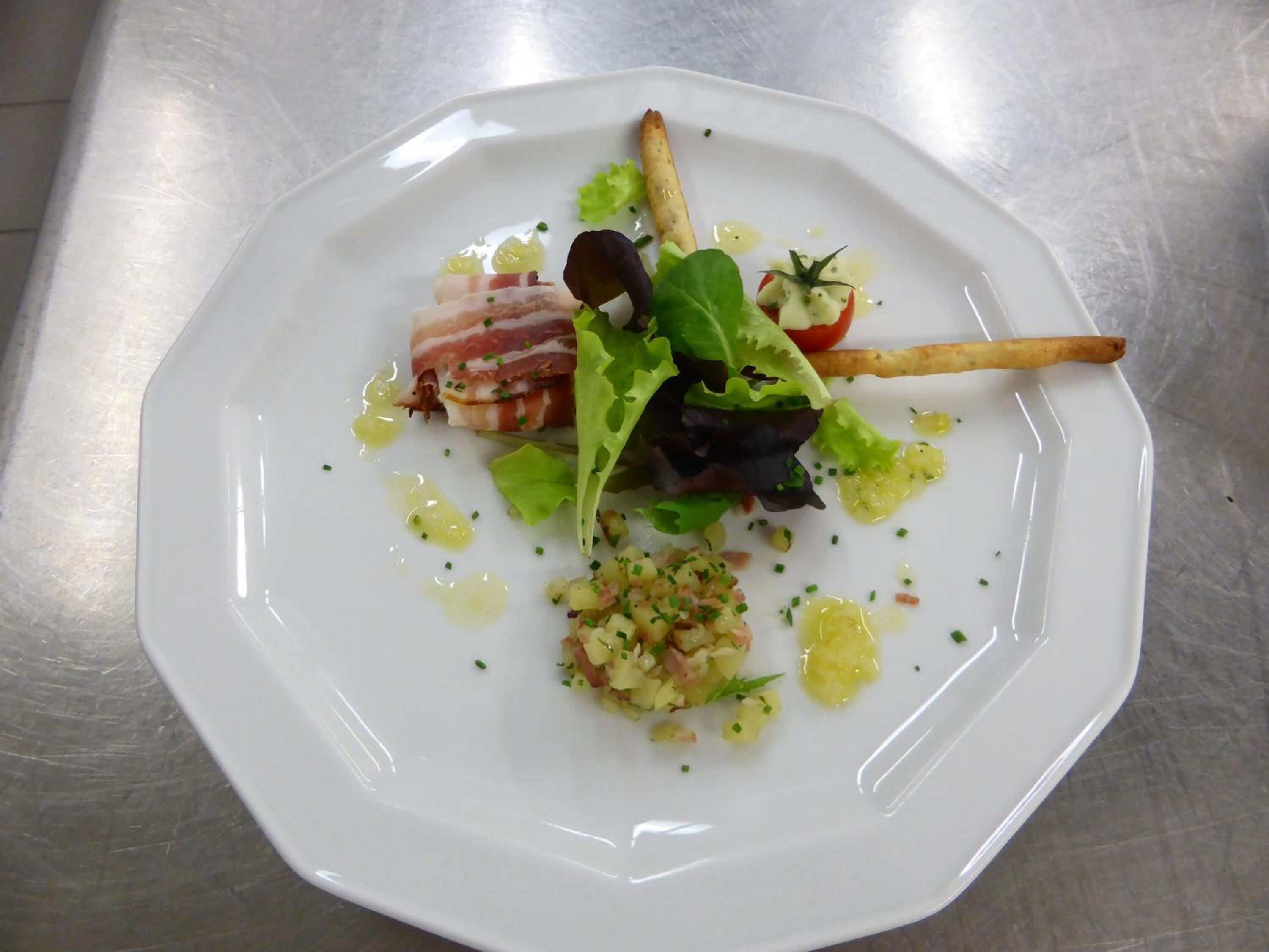 Een wit bord met een salade, spek en een gebakken gerecht op een roestvrijstalen oppervlak.