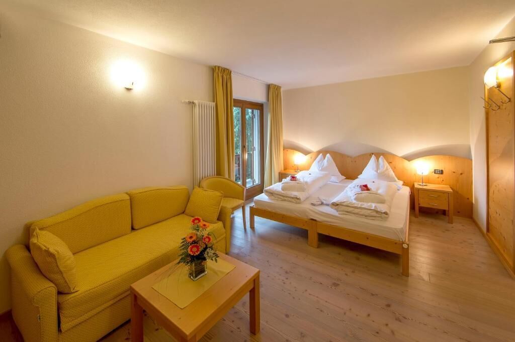 A room with a bed, a yellow sofa, a wooden table with a vase of flowers, and a window with curtains.