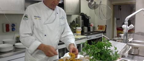 Un chef en una cocina profesional con electrodomésticos de acero inoxidable, diversos utensilios de cocina y una planta de hierbas en maceta.