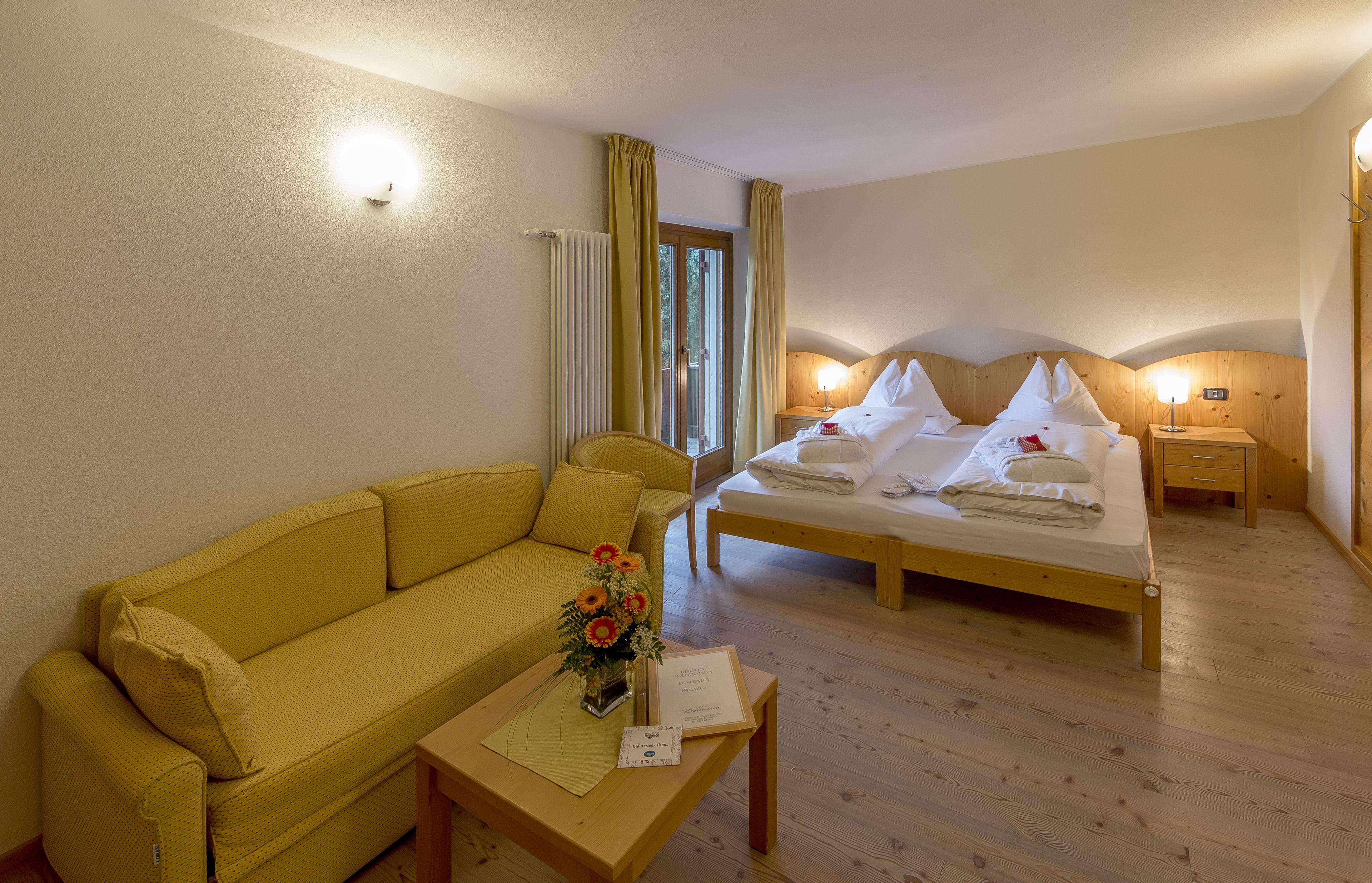 Una habitación de hotel con sofá amarillo, cama de madera, mesita de noche y mesilla con lámpara.