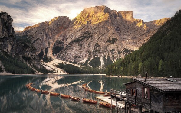 Una cabaña de madera con un muelle que se adentra en un lago tranquilo, rodeada de montañas escarpadas y un bosque denso.