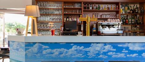 A bar area with a mural of a sky and ocean, shelves of liquor bottles, a coffee machine, and a TV.