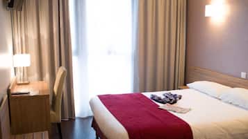 Quarto de hotel com uma cama, uma mesa com abajur, uma cadeira e uma janela com cortinas.