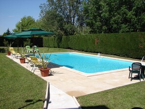 Une piscine rectangulaire avec une terrasse carrelée, entourée d’une pelouse bien entretenue et d’une haie.