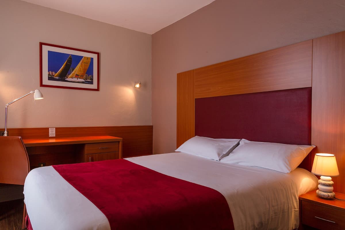Une chambre d’hôtel avec un lit, un bureau, une chaise, une table de chevet, une lampe et un tableau encadré au mur.