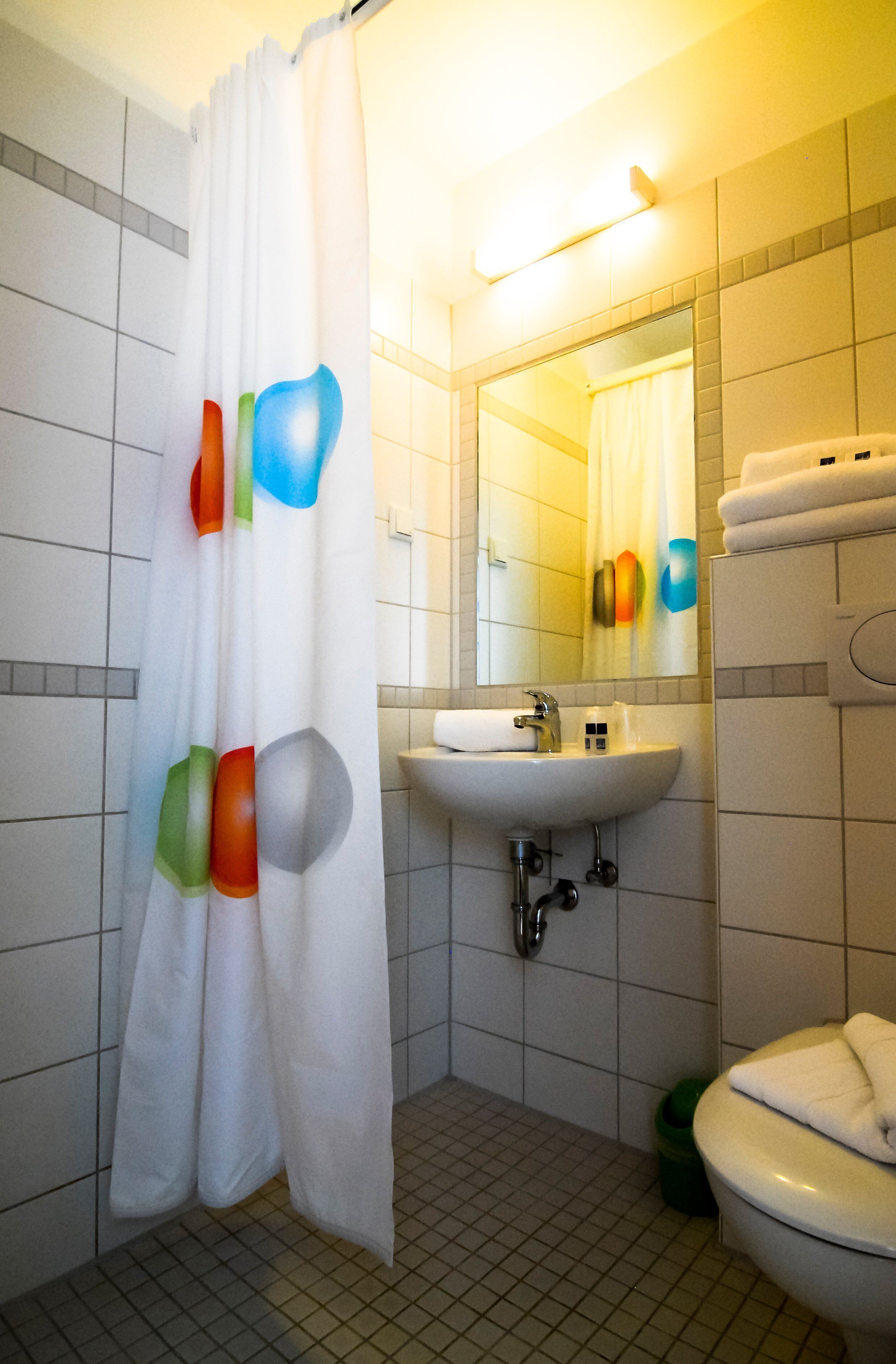 Un bagno con un lavandino bianco, uno specchio, un water e una tenda da doccia con forme colorate.
