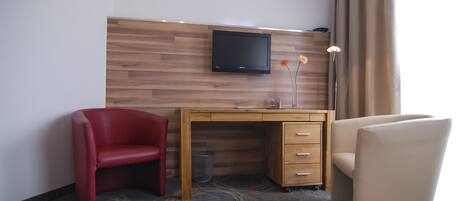 Habitación de hotel con un escritorio de madera, un televisor, una silla roja, una silla color beige y un florero con flores.