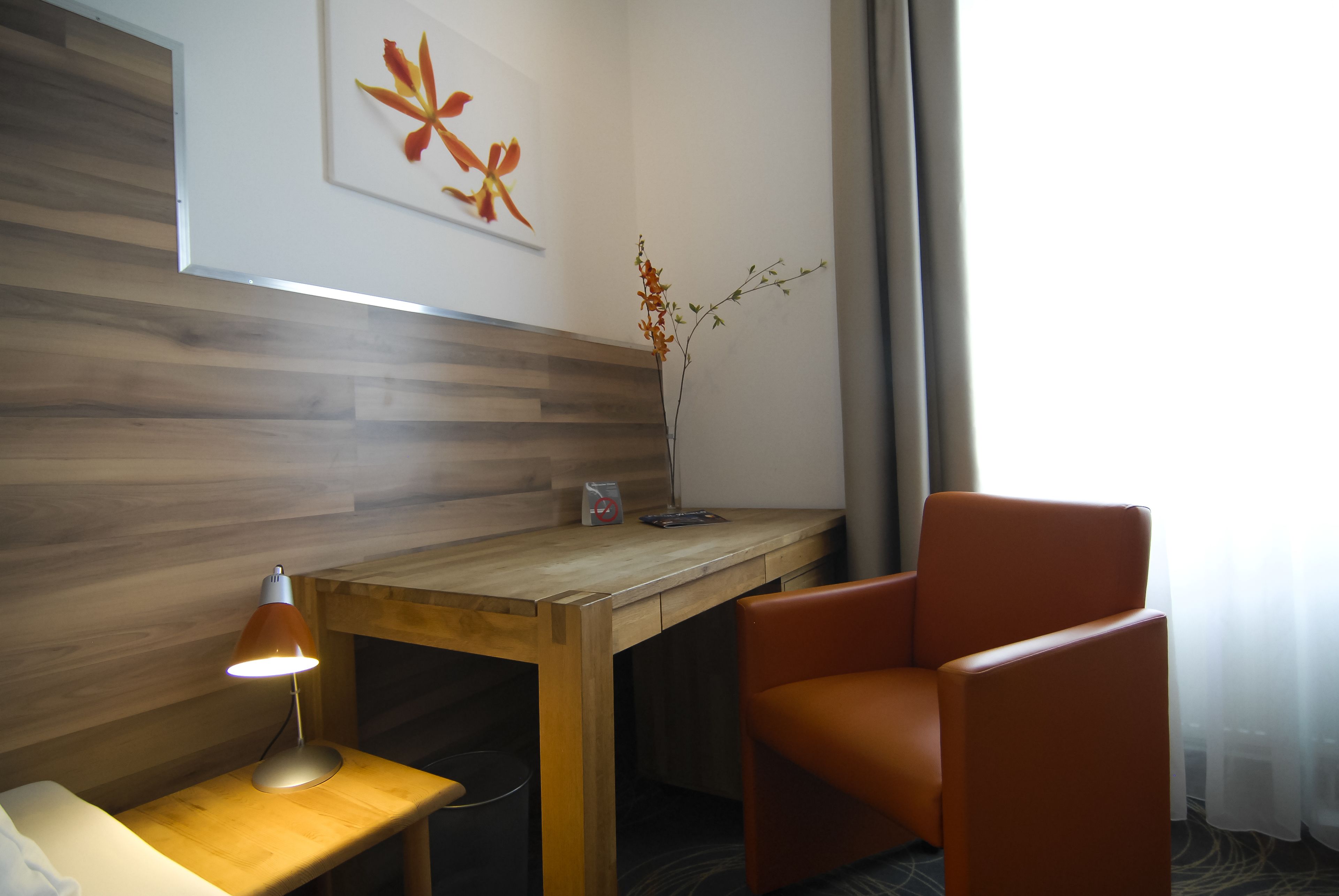 Quarto de hotel com escrivaninha de madeira, uma cadeira laranja, luminária de mesa e um vaso com flores.
