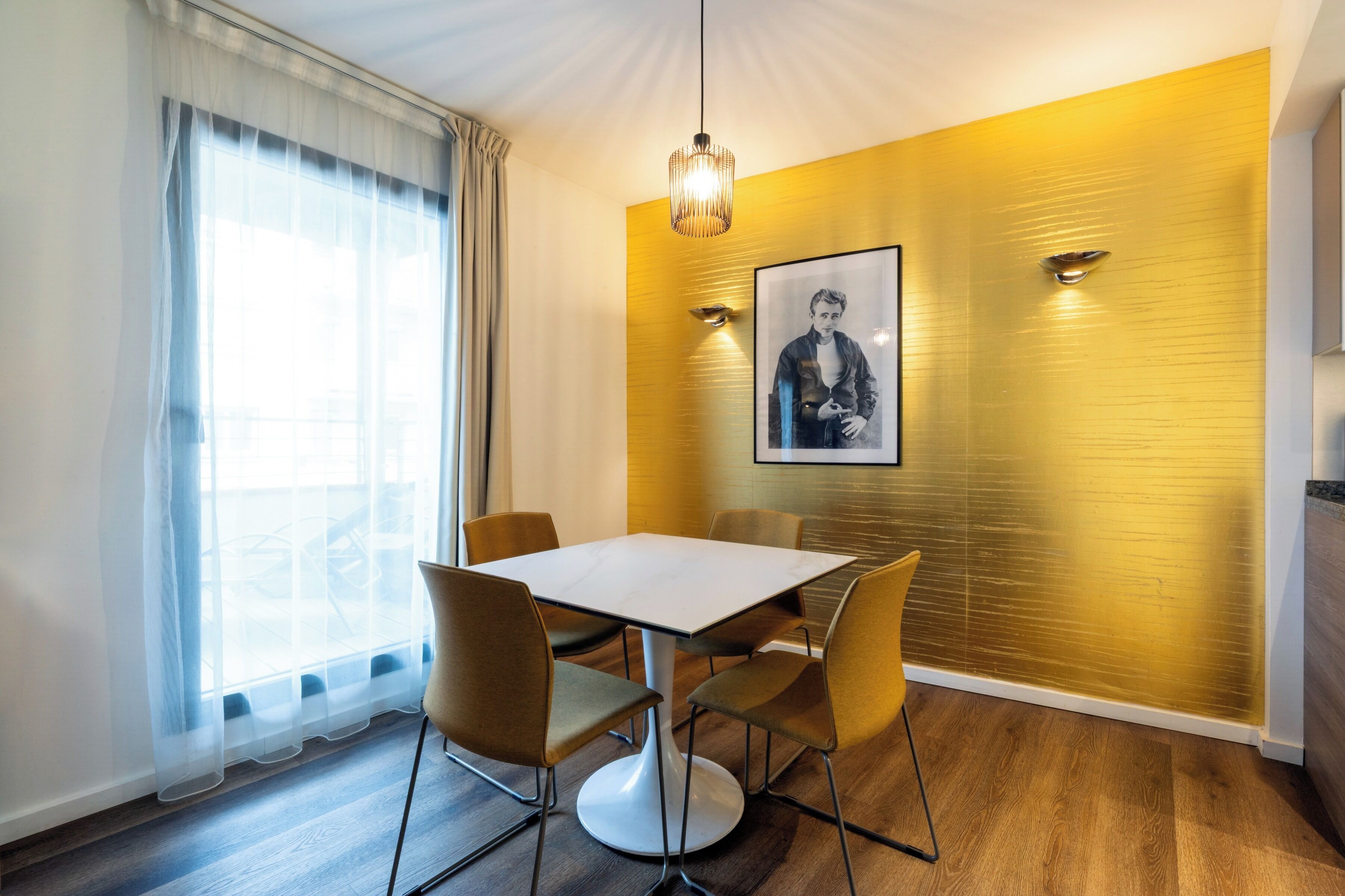A dining area with a table and chairs, a pendant light, a framed picture on the wall, and a window with sheer curtains.