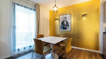A dining area with a table and chairs, a pendant light, a framed picture on the wall, and a window with sheer curtains.