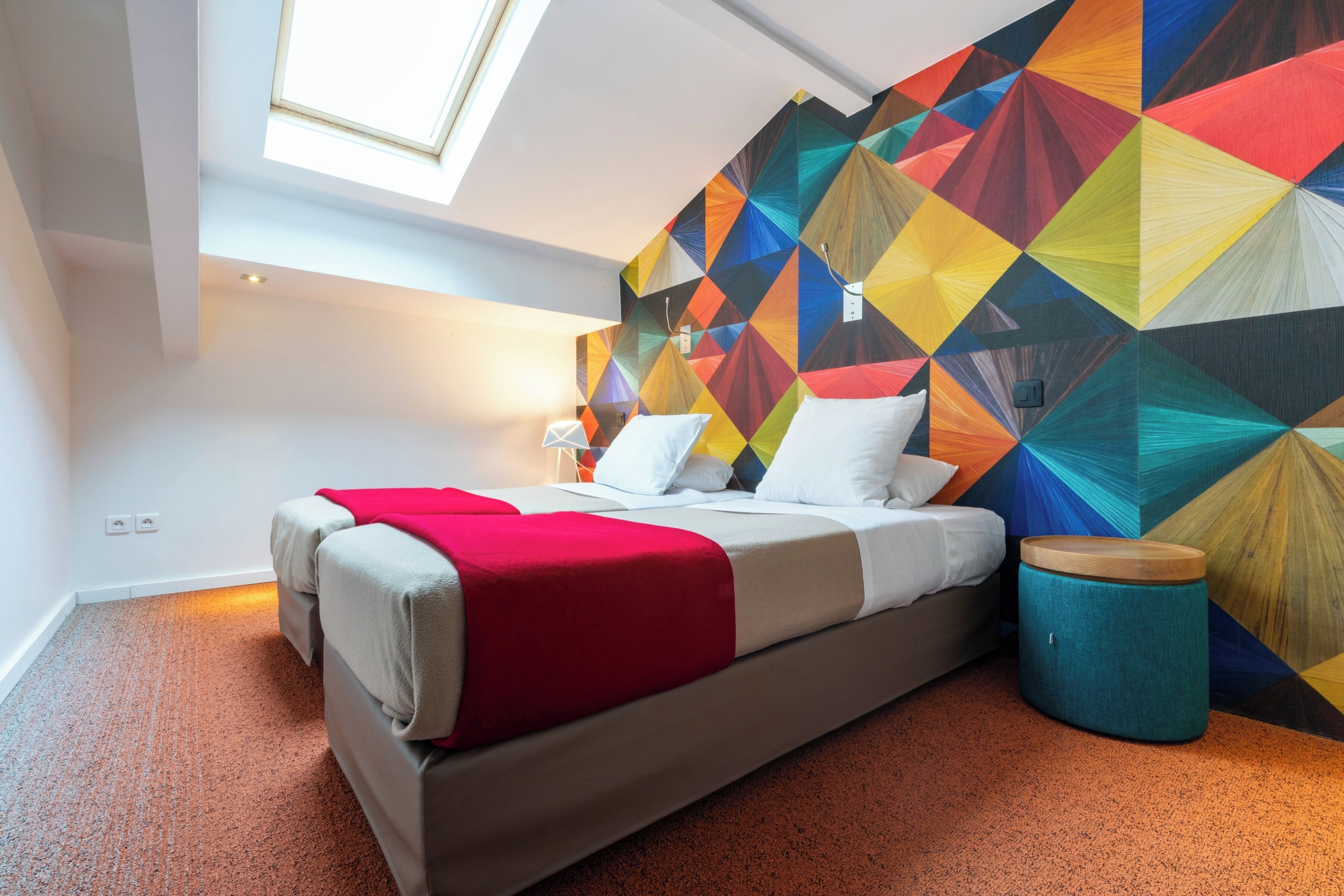 Een moderne slaapkamer met een wand in een geometrisch patroon, een bed met een rode deken en een dakvenster.