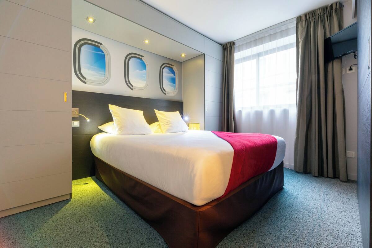 A hotel room with a large bed, a headboard with circular windows, a red throw blanket, and a window with curtains.
