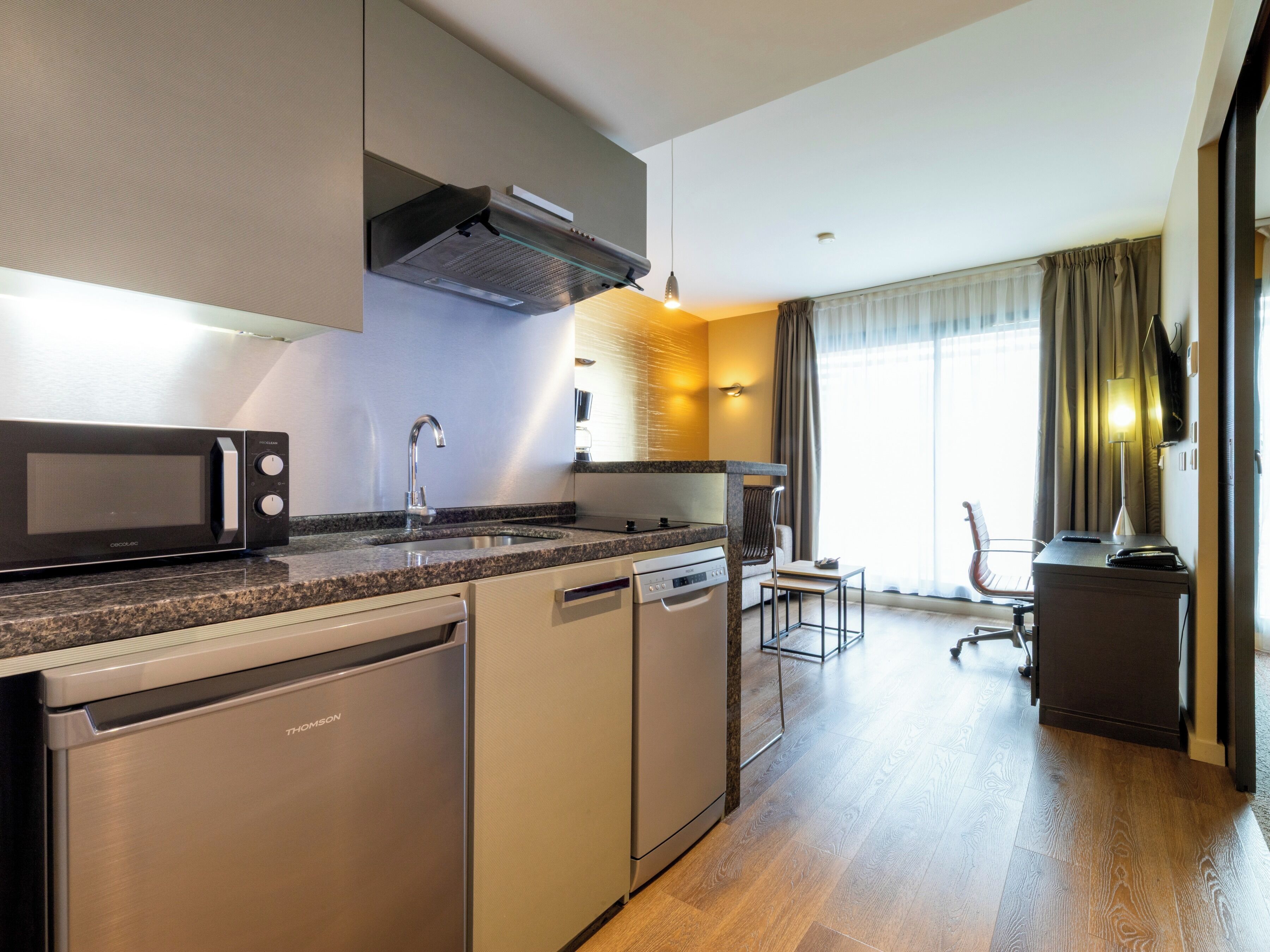 Una cocina moderna con microondas, lavavajillas y estufa integrados. Hay una zona de comedor con una silla y una mesa, además de un gran ventanal con cortinas.