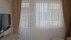 Habitación de hotel con televisor sobre un soporte, una mesita pequeña y una ventana con cortinas translúcidas.