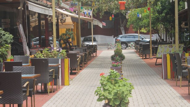 Una fila de mesas y sillas al aire libre sobre un camino pavimentado, con plantas en macetas en el centro.