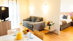 A modern hotel room with a sofa, a small table with breakfast items, a TV, and a bed with a nightstand.