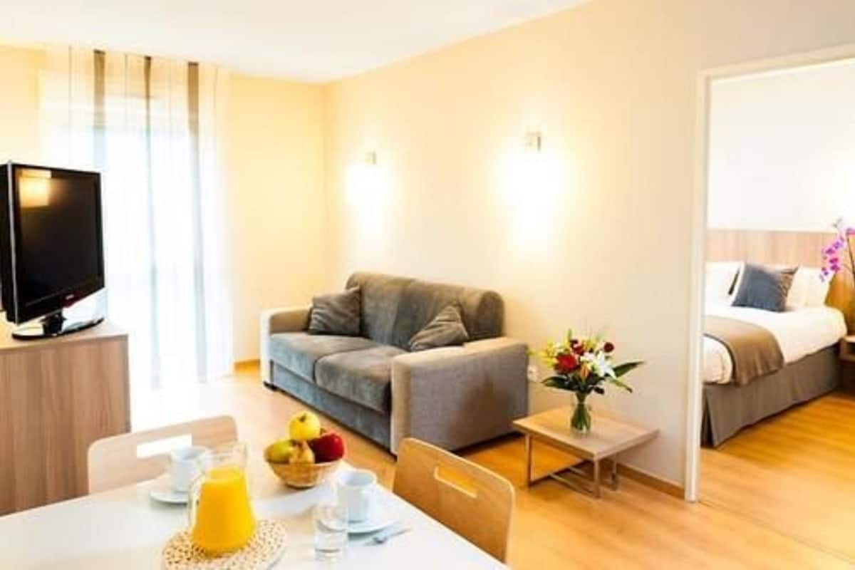 Una habitación de hotel moderna con sofá, una pequeña mesa con productos para el desayuno, televisión y cama con mesita de noche.