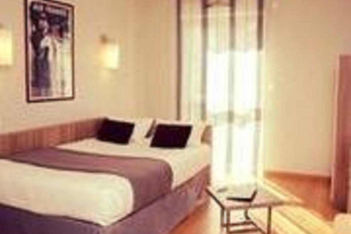 Una habitación de hotel con una cama, una mesita de noche, una ventana con cortinas y un aplique de pared.