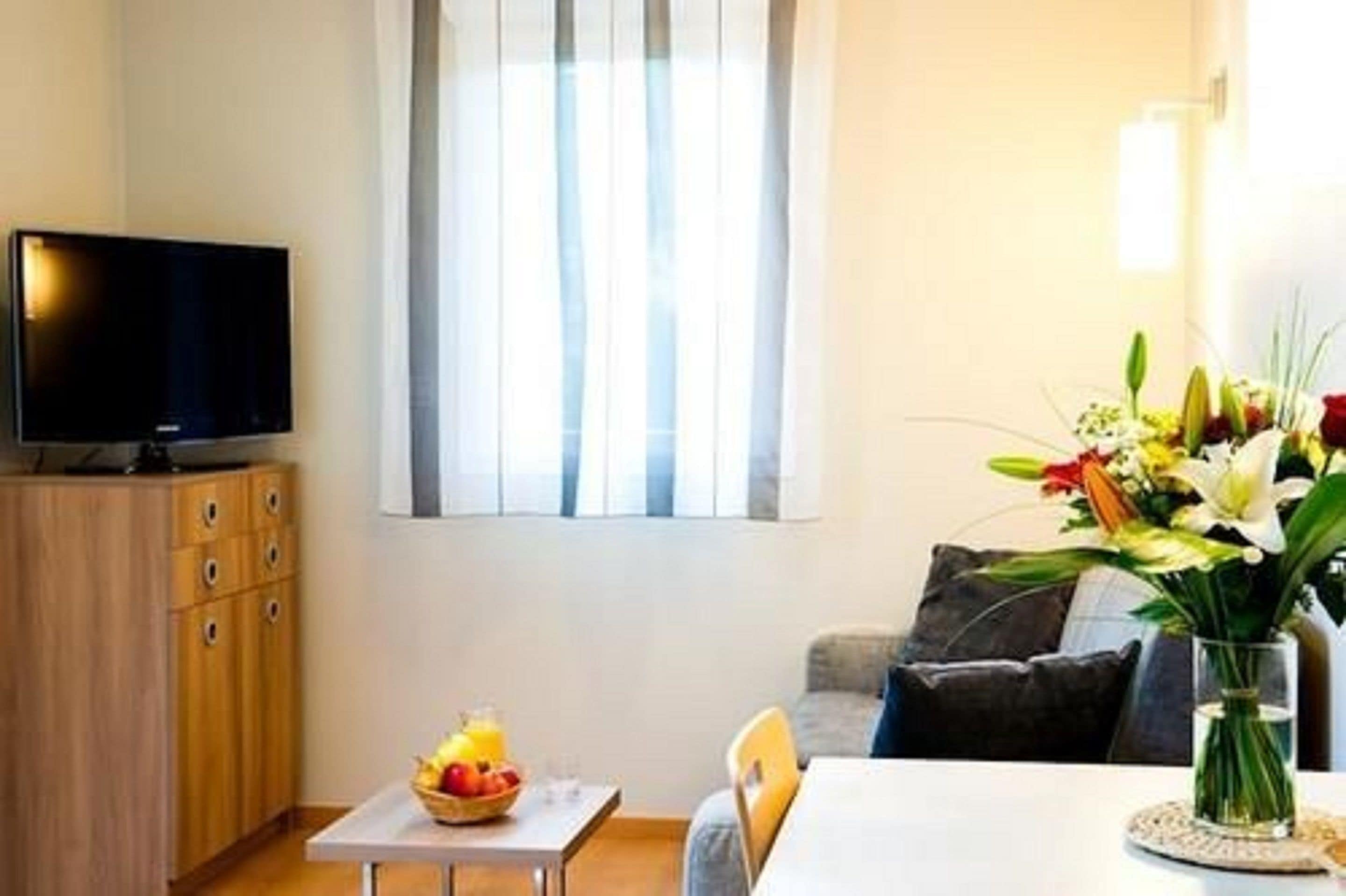 Una sala de estar moderna con un televisor de pantalla plana, una mesa de comedor con frutero, un jarrón con flores y un sofá con cojines.