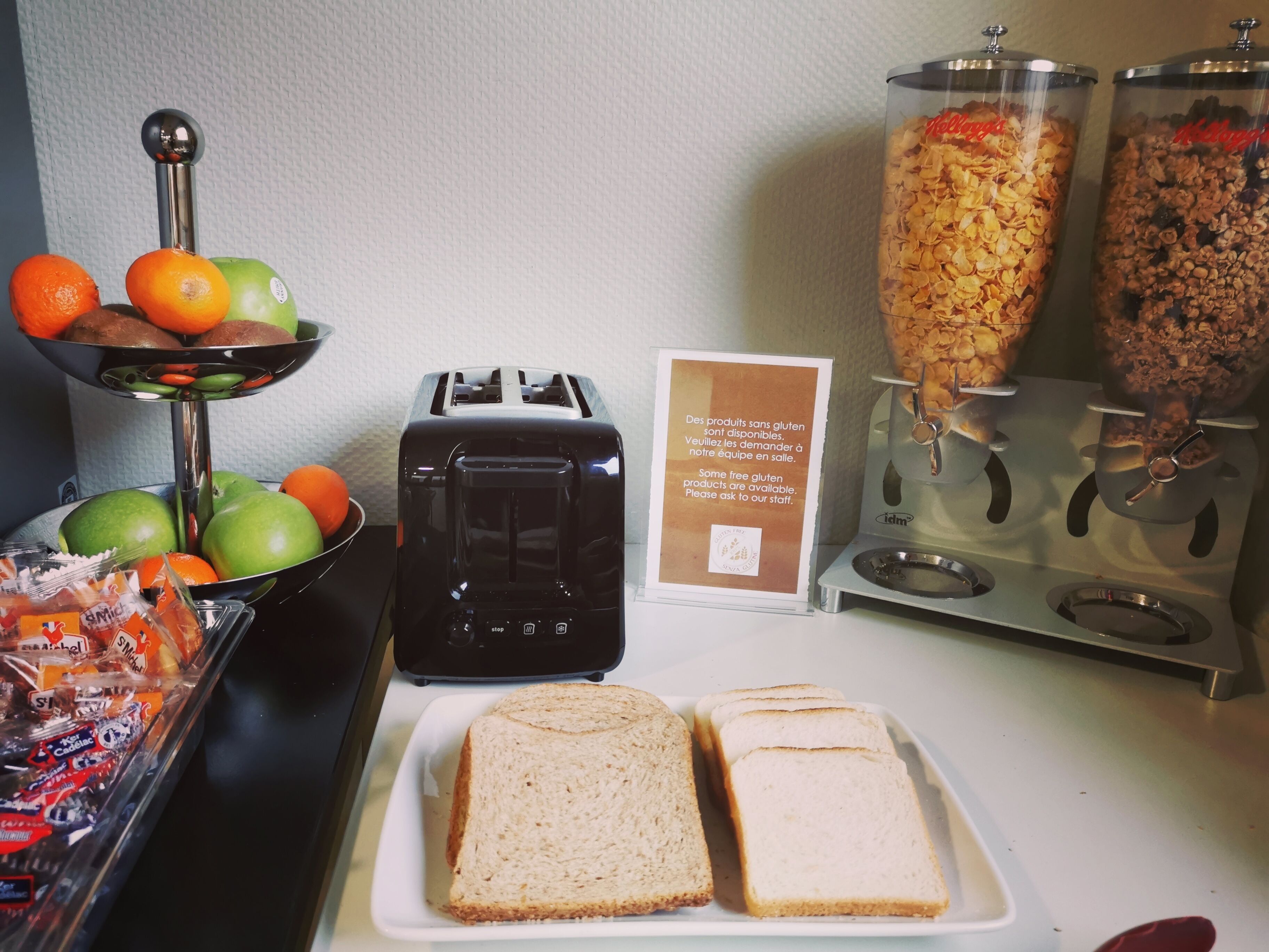 A breakfast buffet with a toaster, a bowl of fruits, and cereal dispensers.