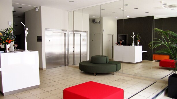 A modern lobby with a reception desk, a green sofa, a red ottoman, and a large mirror.