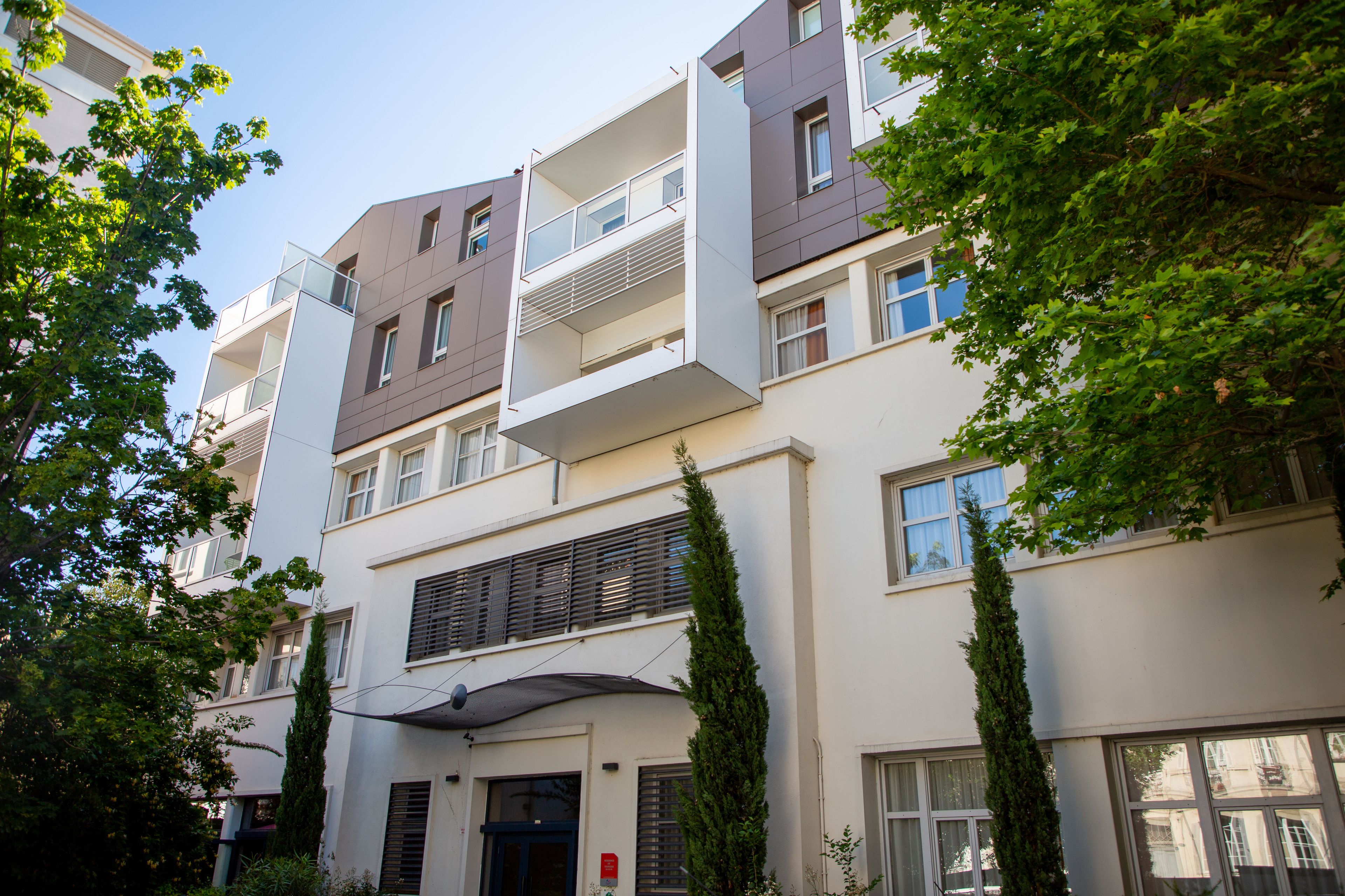 Un edificio residencial de varios pisos con balcones, una entrada central y vegetación circundante.