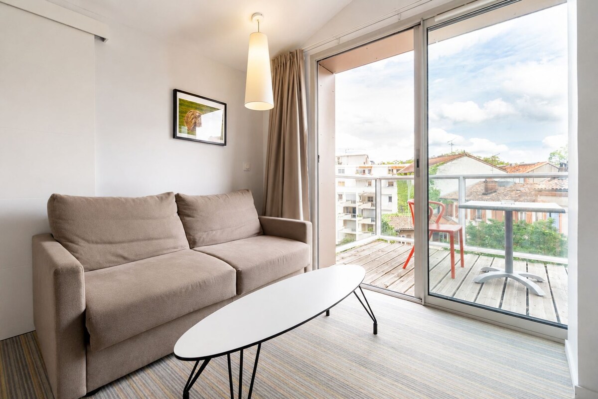 Una habitación moderna con sofá, una mesita, un cuadro en la pared y un balcón con vistas a edificios.