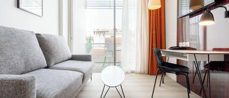 Una sala moderna con un sofá gris, una mesita redonda blanca, una silla negra, una mesa de comedor de madera y un ventanal con cortinas.