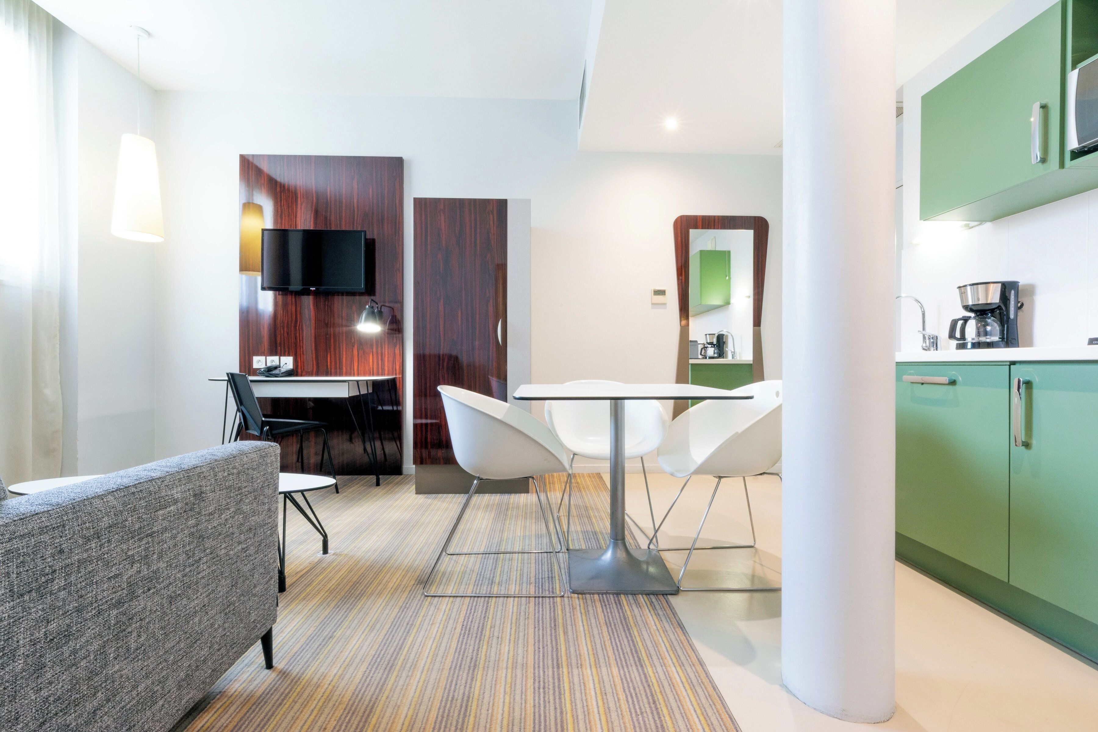 A modern hotel room with a dining area, a TV mounted on a wooden panel, a green kitchen, and a white sofa.