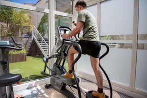 Una persona usando una bicicleta estática en una habitación con ventanas amplias, vista a un patio trasero con terraza de madera y escaleras, y un piso con diseño.