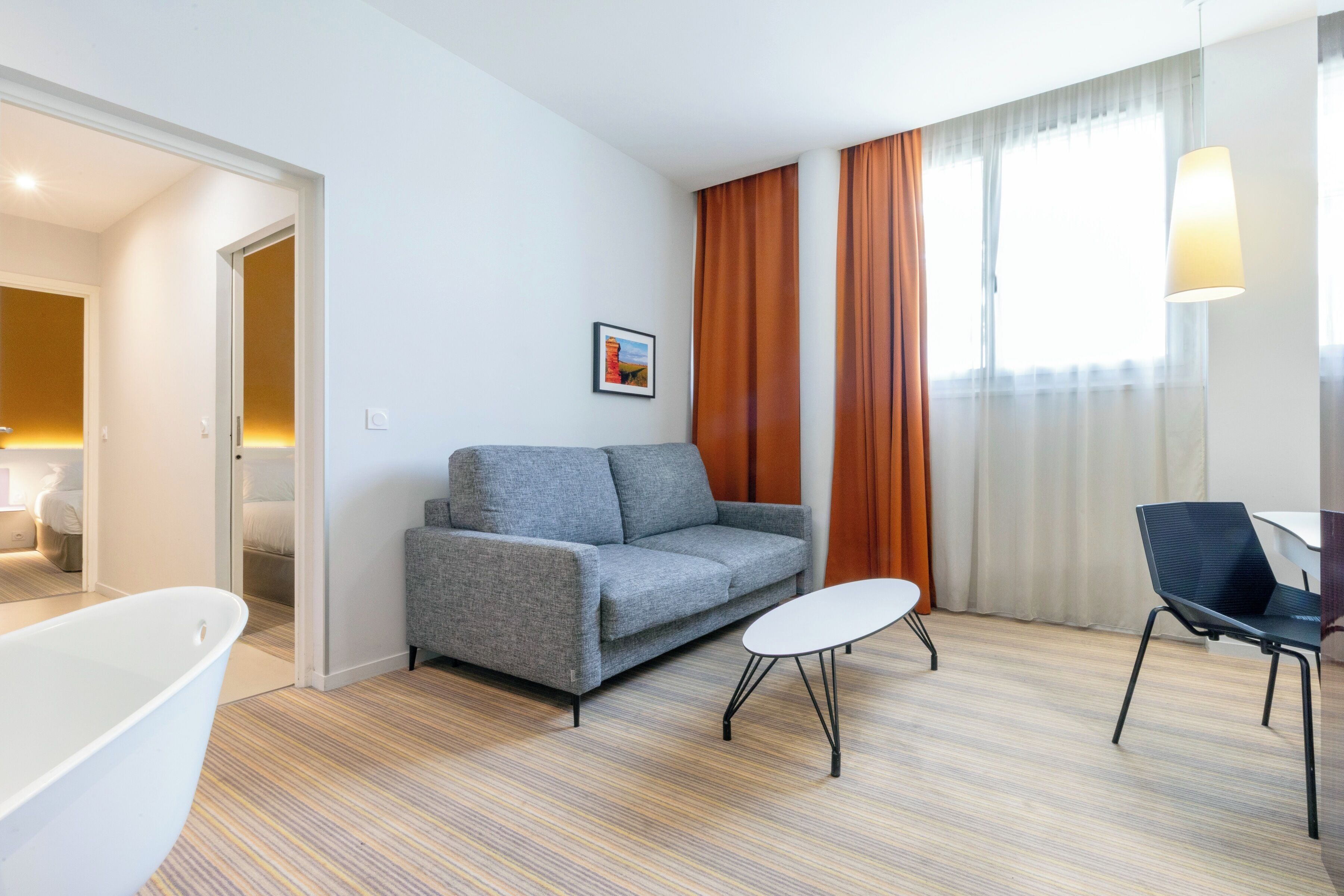 A modern hotel room with a grey sofa, a small round table, a black chair, a white bathtub, and a large window with curtains.