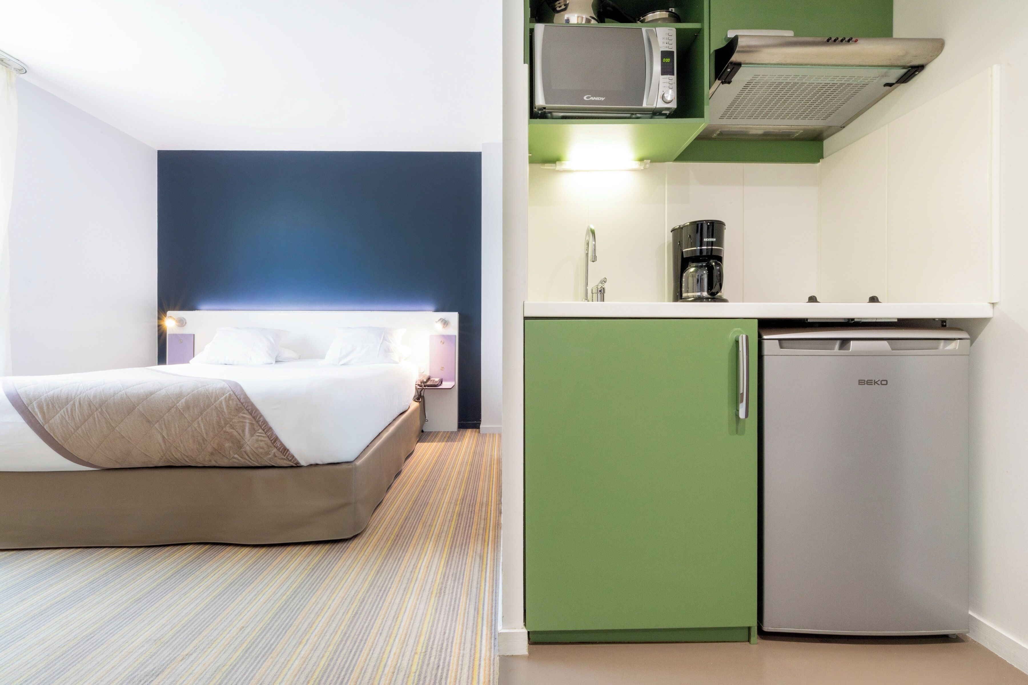 Habitació d'hotel amb llit, cuineta equipada amb microones, cafetera i nevera.