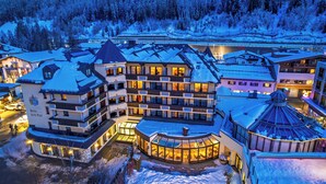 Um hotel de vários andares com um telhado coberto de neve, uma estufa de vidro e uma estação de esqui ao fundo.
