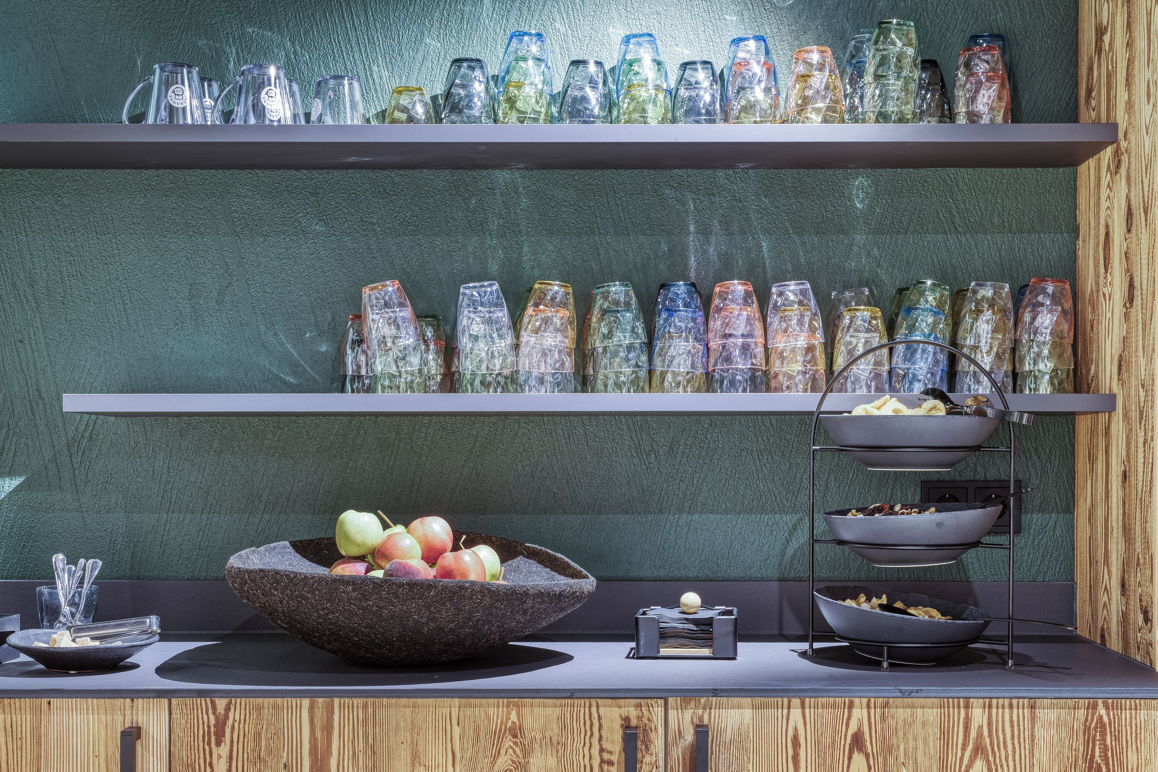 A kitchen counter with a fruit bowl, a metal rack with bowls, and a shelf with glassware.